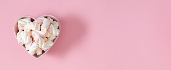 Multicolored marshmallows spirals in a heart-shaped plate on a pink background. Valentine's Day background. Top view. Flat lay. Sweet background, banner.