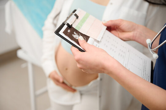 Cropped view of female doctor showing prescription or notes on clipboard to pregnant woman during prenatal care visit in clinic. Pregnant woman receiving prenatal care from doctor. Fetal care
