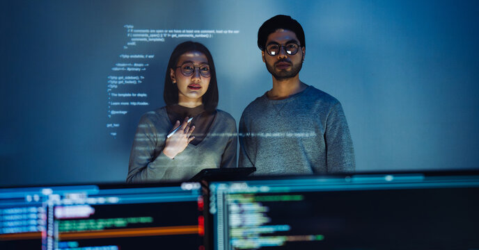 Asian Two software developers engaged in collaborative coding session, with code projected onto the wall and multiple computer screens displaying