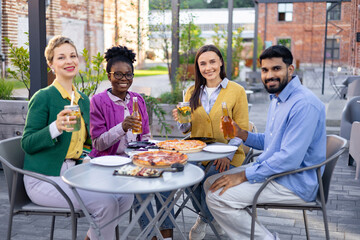 Diverse group of friends enjoying pizza and drinks together outdoors at a cafe, smiling and having...
