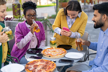Friends enjoying pizza and drinks at an outdoor cafe, capturing moments with a phone and having a...