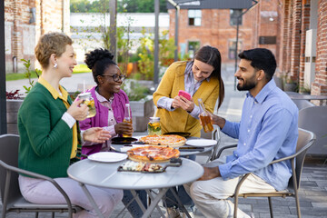 A diverse group of friends enjoys pizza and drinks at an outdoor restaurant, capturing a moment together with a photo.
