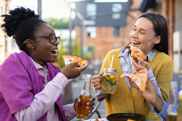 Two cheerful women share pizza and drinks outdoors, enjoying a relaxed meal together, laughing and having fun.