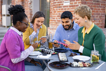 A diverse group of friends are gathered at an outdoor cafe, enjoying food and using their smartphones.