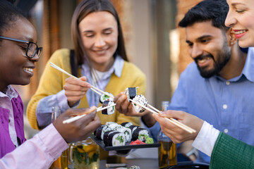 A diverse group of friends enjoys sushi at an outdoor restaurant, using chopsticks and smiling.