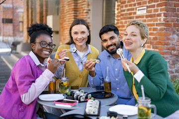 A diverse group of friends enjoys a sushi meal outdoors, smiling and using chopsticks at a table.