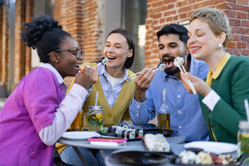 A diverse group of friends enjoys sushi and drinks together at an outdoor restaurant, laughing and having fun.