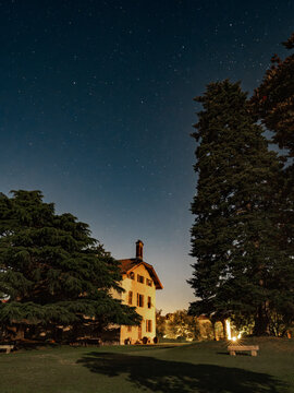 High-resolution nighttime photograph of a rustic Italian villa under a star-filled sky. The scene is framed by tall conifer trees and features warm lighting on the house facade and surrounding garden.