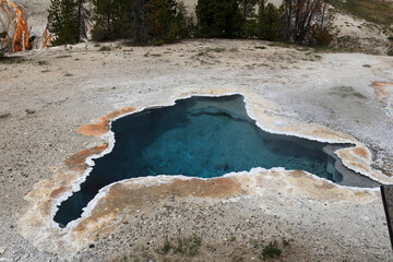 A large thermal spring of bright blue color in Yellowstone. There is gray-dirty soil around the...