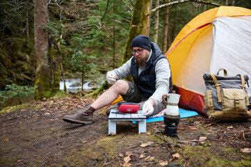 Hiker man with injured leg using first aid kit near the tent in the forest. A stressed trekker...