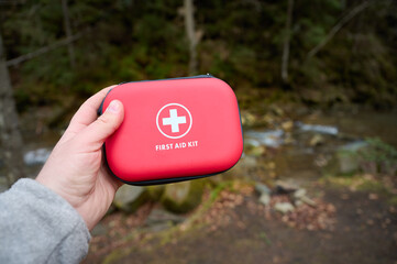 Closeup view of a hand holding a red first aid kit near a camping tent in the forest. First aid...