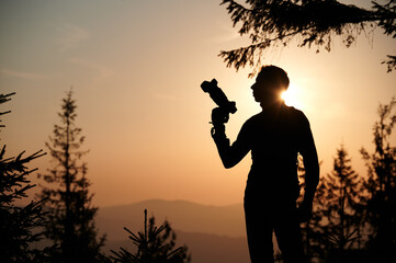 Silhouette of a man holding a camera in the mountains at sunset. Photographer in nature, backlit by...