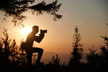 Silhouette of a man holding a camera in the mountains at sunset. Photographer in nature, backlit by...