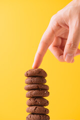 Finger Touching Stack of Chocolate Cookies on Yellow Background, Balanced Cookie Tower with Hand on Top, Stack of Chocolate Cookies Being Touched by a Finger, Hand Reaching for Cookie from a Tall Stac
