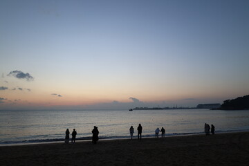 Fototapeta premium People Silhouetted on Beach at Twilight with Calm Ocean and Distant Coastline Under Evening Sky