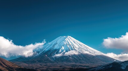 Fototapeta premium Snow-capped mountain under a clear blue sky with clouds floating around the peak.