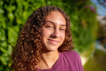 Hispanic teenage girl with curly hair standing confidently in sunlit park, expressing vibrant youth...