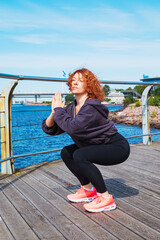 Energetic red-haired curly young woman in sportswear doing squats on river embankment.