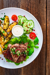 Grilled beef steak with baked potato wedges and fresh vegetables on wooden table	