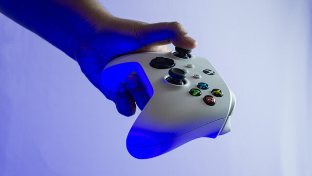 South Kalimantan, Indonesia - 07 14 2025 : White Xbox Series S|X controller held in one hand on white background under blue ambient light, showing minimal modern gaming design