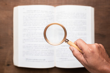 Hand holding magnifying glass over open book on wooden table. Concept of reading, studying,...