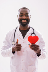 Portrait of smiling black doctor who is holding heart shape pillow and showing thumb up on gray...