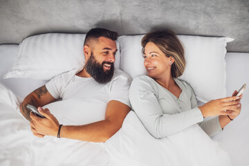 Young happy couple is lying in bed in the morning. They are relaxing and using smartphones.