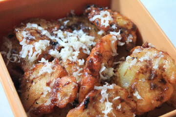 Close-up view of golden-brown fried bananas, topped with grated white cheese and drizzled with a dark sauce, presented in an orange rectangular dish. Related to themes related to comfort food