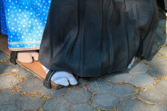 Close-up of individuals wearing traditional Japanese garments and standing on a single, long wooden platform-style footwear, paired with white split-toe tabi socks, as they navigate a paved ground