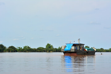 Dark-hulled boat with a blue tarp covering part of its deck, floating on a calm, muddy river,...