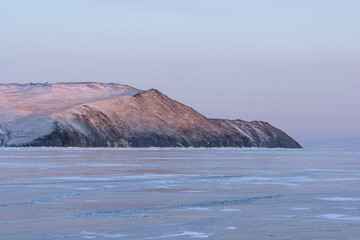 Snow-covered mountain peak rises above frozen lake, reflecting soft pastel colors of dawn, creating a serene winter landscape with tranquil atmosphere and natural beauty