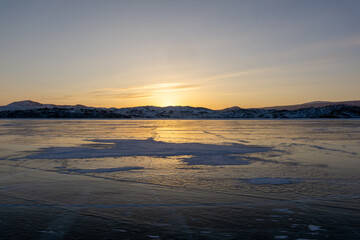 Fototapeta premium Expansive frozen lake reflecting golden sunset hues, with distant snow-capped mountains under a clear sky, creating a serene winter landscape atmosphere