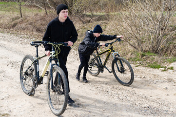 Naklejka premium Two teens walk their bikes down a dirt road, one pushing his bike while the other walks alongside.