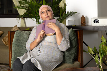 Asian woman recovering from cancer sitting on sofa and reading interesting book as hobby while...