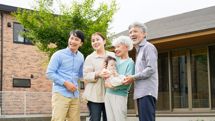 A three-generation family smiling and surrounding a baby in front of their house
