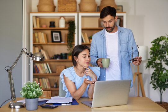 Two individuals are engaged in a discussion over a laptop in a modern home office filled with books and plants while enjoying a relaxing afternoon together.