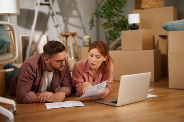 A couple is lying on the floor in a welcoming room, discussing moving details while reviewing paperwork on a laptop. Cardboard boxes are scattered around, indicating the transition to their new home.