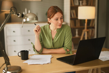 A young woman sits at a wooden desk in a cozy home office, focused on her laptop while jotting down...