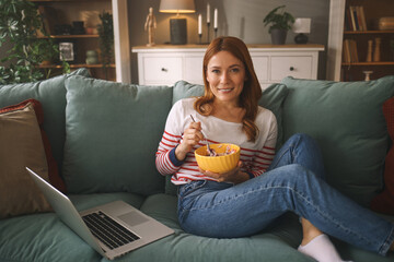 A woman with long red hair sits comfortably on a couch, smiling while eating dessert from a bowl. A laptop rests beside her in a cozy, well-decorated living room.