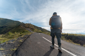 A lone hiker with a backpack walks up an empty mountain road under the sunrise sky, symbolizing...