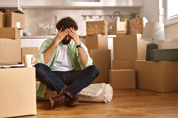 A man sits on the floor with his hands on his head, looking stressed while surrounded by cardboard...