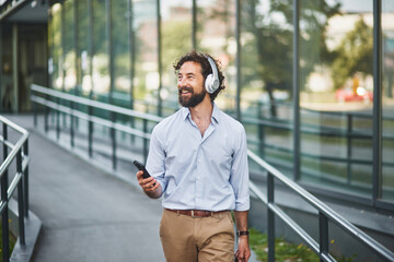 A man dressed in a light blue shirt and beige pants strolls through a glass-enclosed walkway, enjoying music through headphones while using his smartphone in a business district.