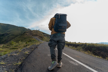 A lone hiker with a backpack walks up an empty mountain road under the sunrise sky, symbolizing...