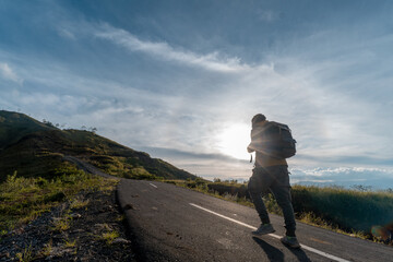A lone hiker with a backpack walks up an empty mountain road under the sunrise sky, symbolizing...