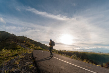 A lone hiker with a backpack walks up an empty mountain road under the sunrise sky, symbolizing...