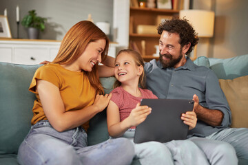 A family of three shares smiles and laughter while sitting on a couch in their living room. The parents engage with their daughter as she plays with a tablet, creating joyful moments.