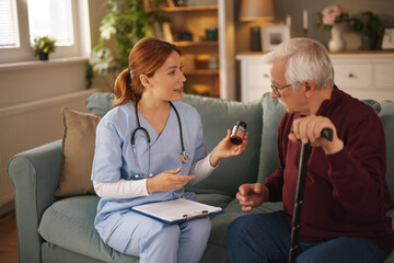 A female nurse in blue scrubs is sitting with a senior man on a couch. She is explaining how to...