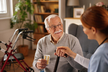 An elderly Caucasian man sits in his living room while a red-haired female nurse hands him a cane...