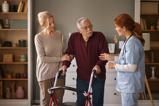 A red-haired female nurse wearing scrubs speaks with an elderly man who is using a walker, while his wife stands beside him holding his arm in a living room setting during the day.