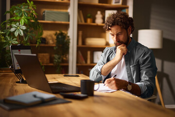A man is seated at a wooden table in his home office, thoughtfully writing notes while surrounded...